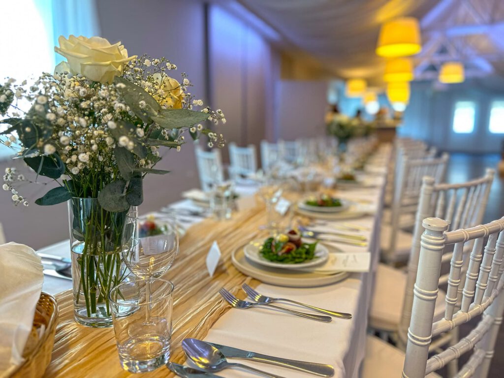 City Club & Garden: Detailný pohľad na slávnostne prestretý dlhý stôl s bielym obrusom a zlatou stredovou štólou. V popredí je sklenená váza s kyticou bielych ruží a gypsomilky, vedľa ktorej sú pripravené taniere s predjedlom a vyleštený príbor.