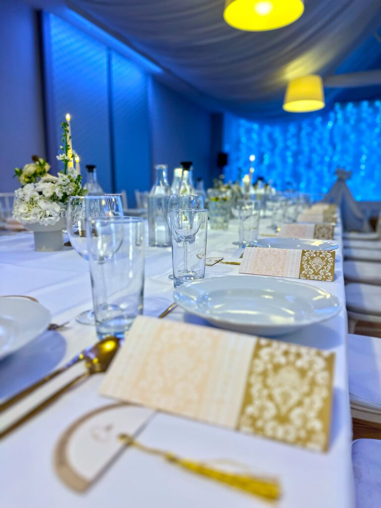 City Club & Garden: Detail prestierania v čistom bielom štýle so zlatými akcentmi. Na stole sú ozdobné menu karty so zlatým vzorom, priehľadné fľaše s vodou, poháre a biela kvetinová dekorácia. Pozadie je ladené do modrého náladového osvetlenia.