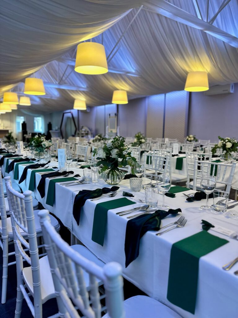 City Club & Garden: Pohľad na usporiadanie stolov v tvare písmena U. Biela téma sýto doplnená zelenými textilnými prvkami a nízkymi sklenenými vázami s bielymi kvetmi. Celý priestor pôsobí vzdušne a slávnostne vďaka baldachýnu a visiacim lampám.