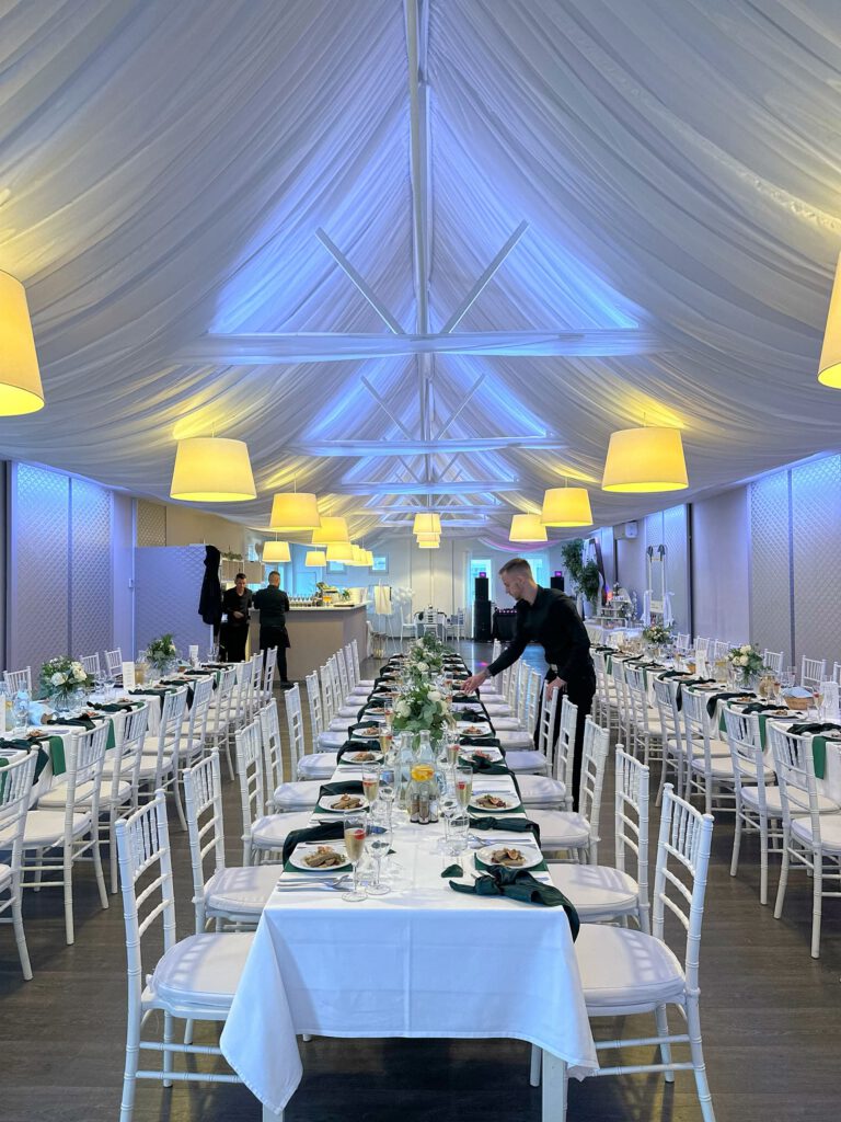 City Club & Garden: Široký záber na symetricky usporiadanú svadobnú sálu, kde čašník dokončuje posledné úpravy na dlhých stoloch. Miestnosť je osvetlená teplými lampami a modrým podsvietením, čo vytvára moderný kontrast.