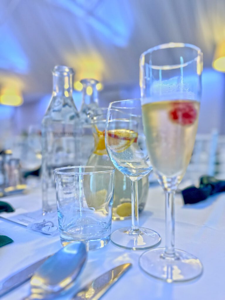 City Club & Garden: Umelecký makro záber na pohár sektu s malinou v popredí. V pozadí sú rozmazané karafy s vodou a citrusmi, zachytávajúce sviežosť podávaných nápojov.