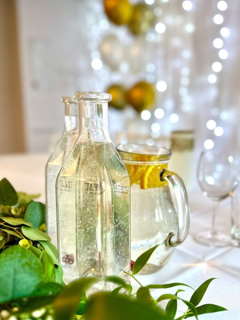 City Club & Garden: Detailný záber na sklenené karafy s vodou a džbán s citrónmi. V popredí je časť zelenej girlandy, v pozadí svetelná stena s balónmi.
