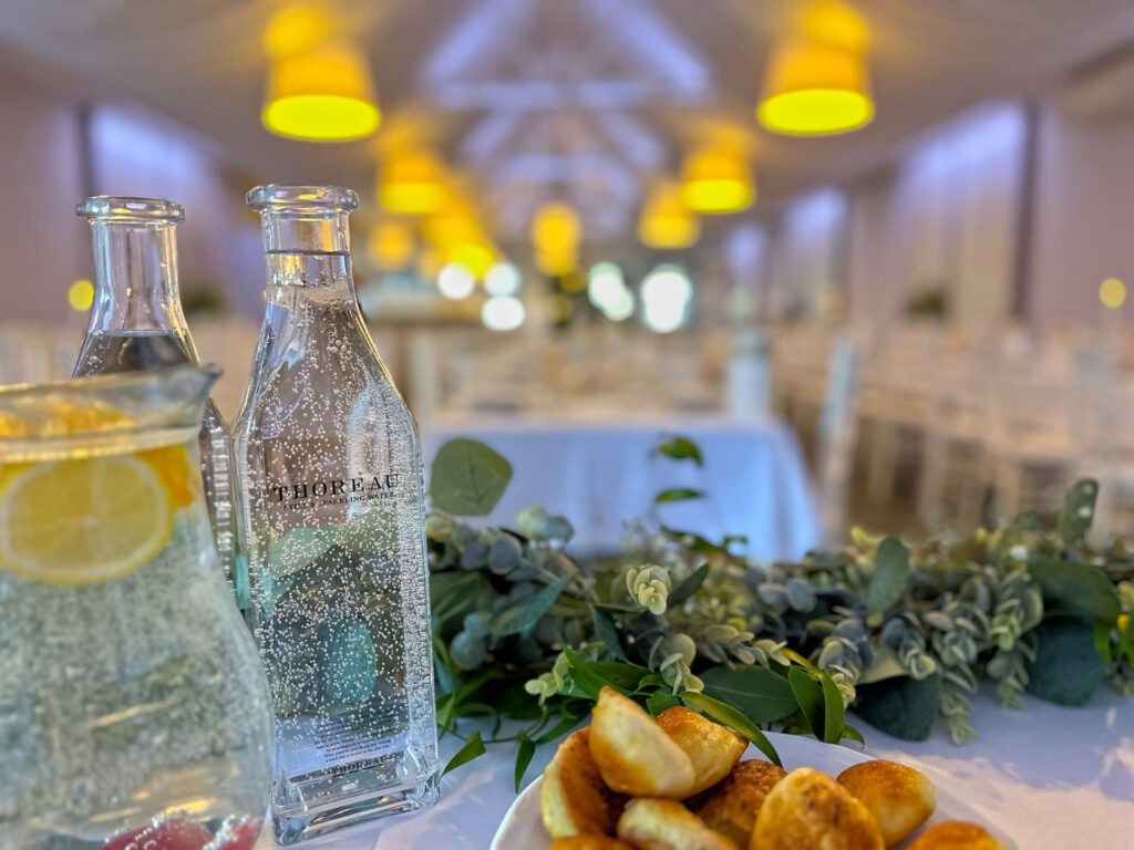 City Club & Garden: Svieži záber na fľašu pramenitej vody značky Thoreau a tanier s pečivom na svadobnom stole, s rozmazaným pohľadom do hĺbky slávnostnej sály.