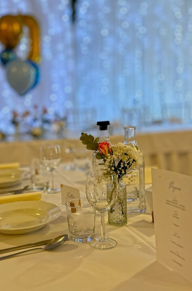 City Club & Garden: Detailné prestieranie s bielym tanierom, príborom a pohármi. V strede stola je sklenená váza s jednou ružou a drobnými bielymi kvetmi, vedľa ktorej stoja fľaše s vodou na pozadí svietiacej steny.