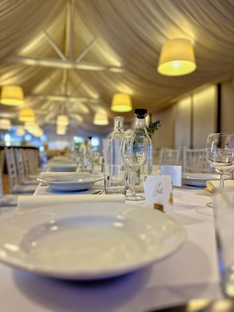 City Club & Garden: Perspektívny pohľad pozdĺž dlhého stola v moderne osvetlenej sále s bielym nariaseným stropom. Stôl je pripravený pre hostí s bielymi taniermi, pohármi na víno a fľašami s vodou.