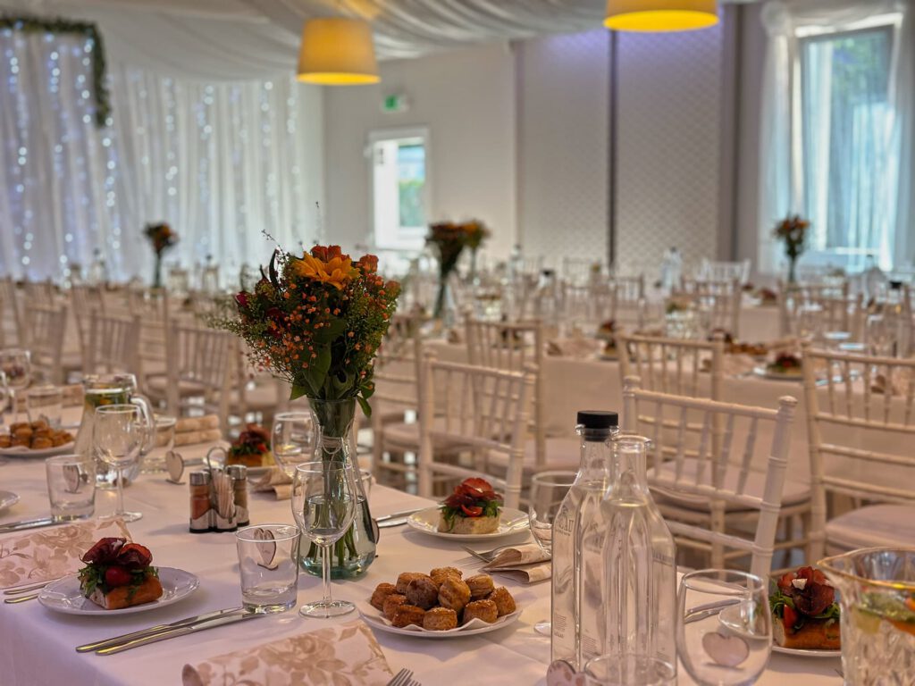 City Club & Garden: Atmosféra slávnostne pripravenej sály s bielymi stoličkami a stolmi. Na stole sú taniere s predjedlom, misky s pečivom, fľaše s vodou a vázy s kvetmi, ktoré ladia s čistým dizajnom priestoru.