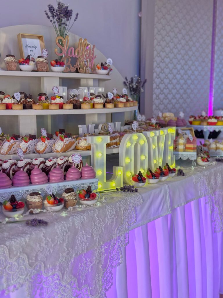 City Club & Garden: Bohatý sladký bar (Candy bar) podsvietený fialovým svetlom. Na bielych stupienkoch sú zoradené rôzne dezerty, ovocné poháre a koláčiky, doplnené veľkým svietiacim nápisom „LOVE“.