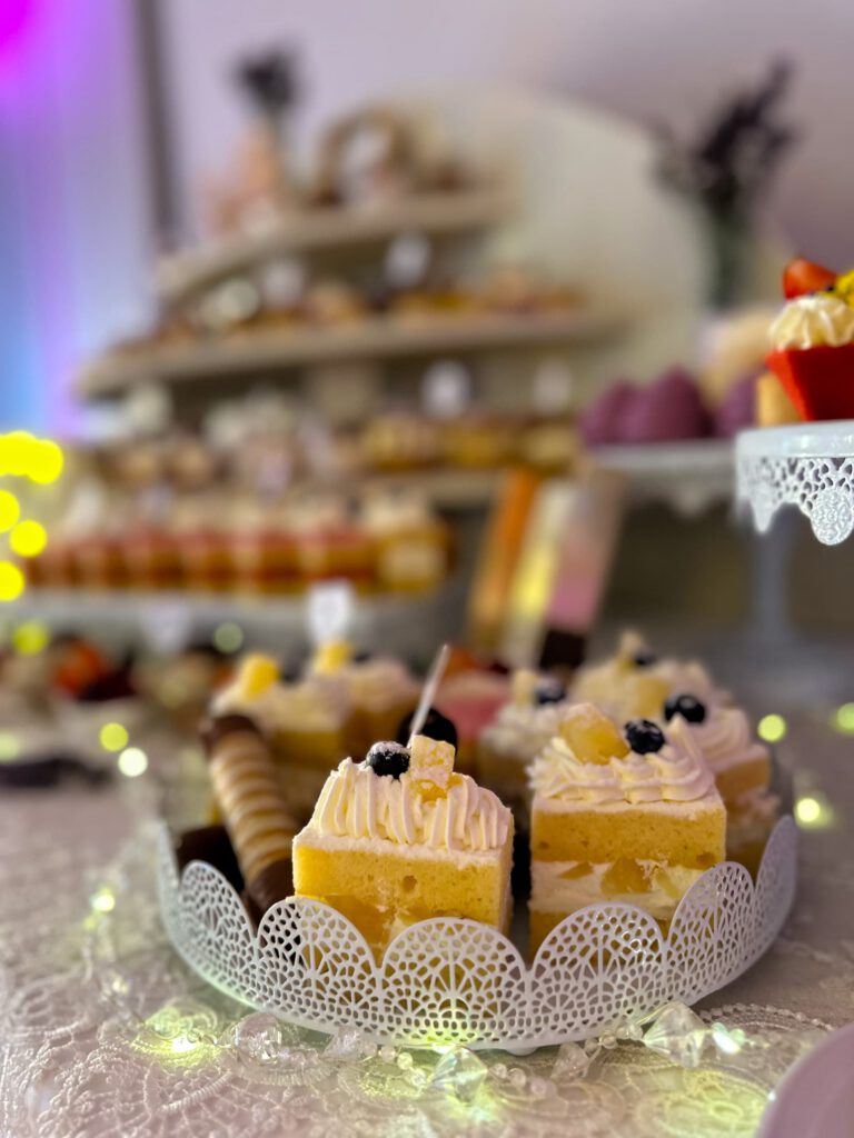 City Club & Garden: Detailný záber na jemný krémový zákusok s ovocím a šľahačkou umiestnený na ozdobnom bielom podnose. V pozadí sa v rozostrení črtá bohato naložený sladký bar so svetielkami.