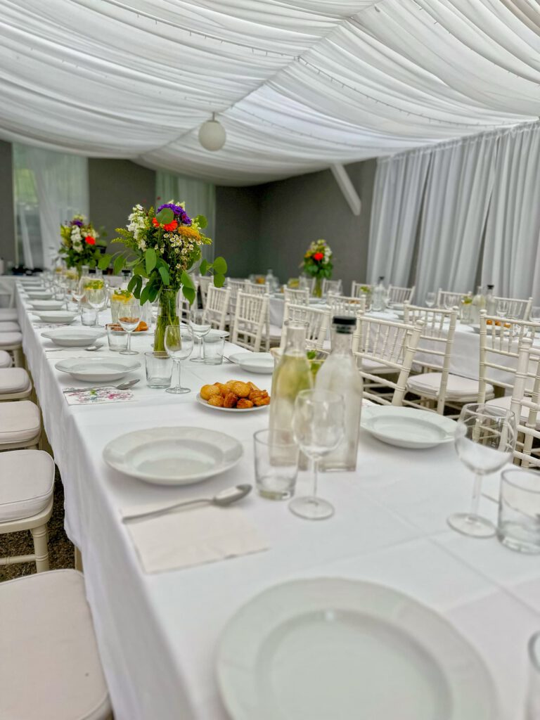 City Club & Garden: Dlhé biele stoly usporiadané pod stanom s nariaseným stropom. Výzdobu tvoria vysoké vázy s pestrofarebnými poľnými kvetmi, taniere s pečivom a džbány s vodou.