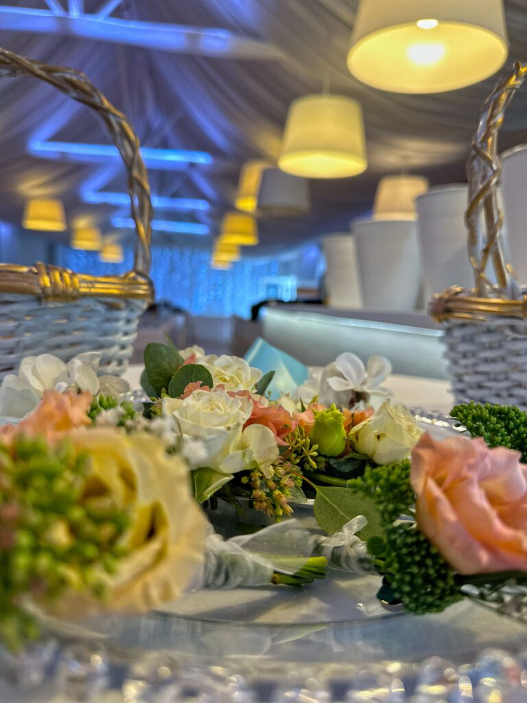 City Club & Garden: Detailný záber na kvetinové pierka (boutonnières) z drobných ruží pripravené na tácke. V pozadí sú vidieť prútené košíky a rozostrený interiér osvetlenej sály.