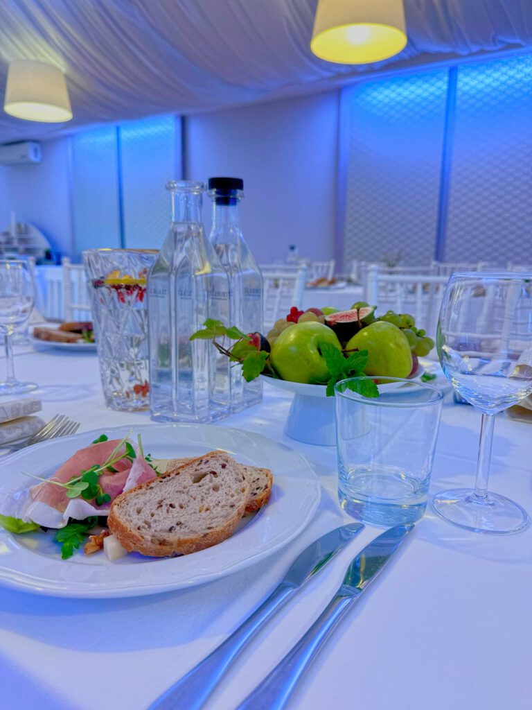 City Club & Garden: Detail prestierania s predjedlom pozostávajúcim zo šunky a čerstvého pečiva. Na stole sa nachádza aj dizajnová misa s ovocím (jablká, figy, hrozno), džbán s citrusovou vodou a čisté poháre na víno pod modrým náladovým osvetlením.