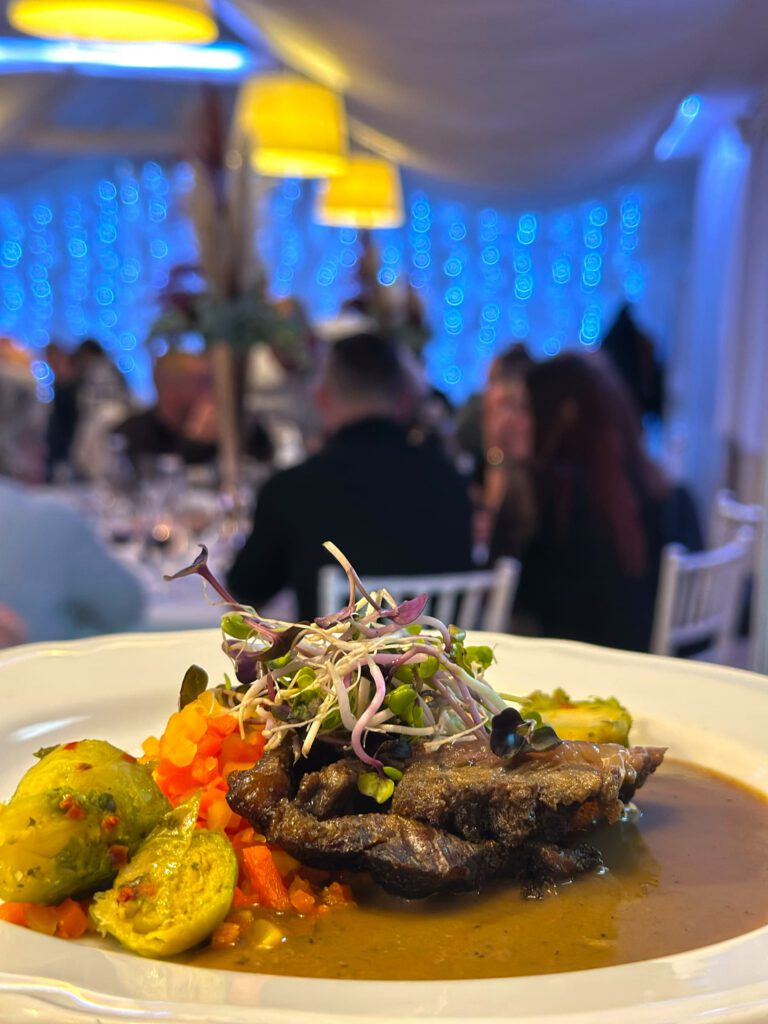 City Club & Garden: Detailný záber na hlavné jedlo – šťavnaté pečené mäso s omáčkou, podávané s pečeným ružičkovým kelom, mrkvou a dozdobené čerstvými klíčkami. V pozadí je vidieť atmosféru prebiehajúcej oslavy pri modrej svetelnej stene.